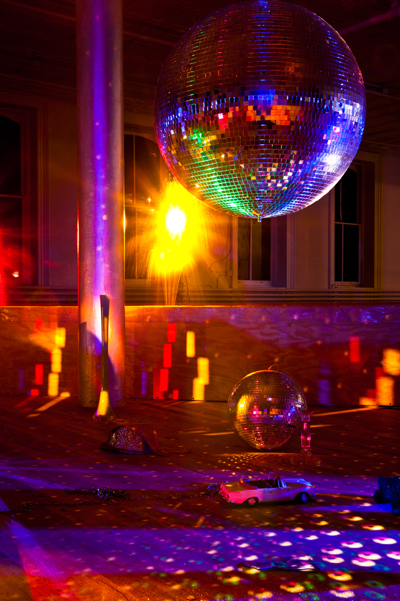 A disco ball reflects multicolored light spots across a room lit by pink and yellow hues. On the floor sit a second disco ball, a silver glittery hat, a silver chain, and a white convertable toy car.
