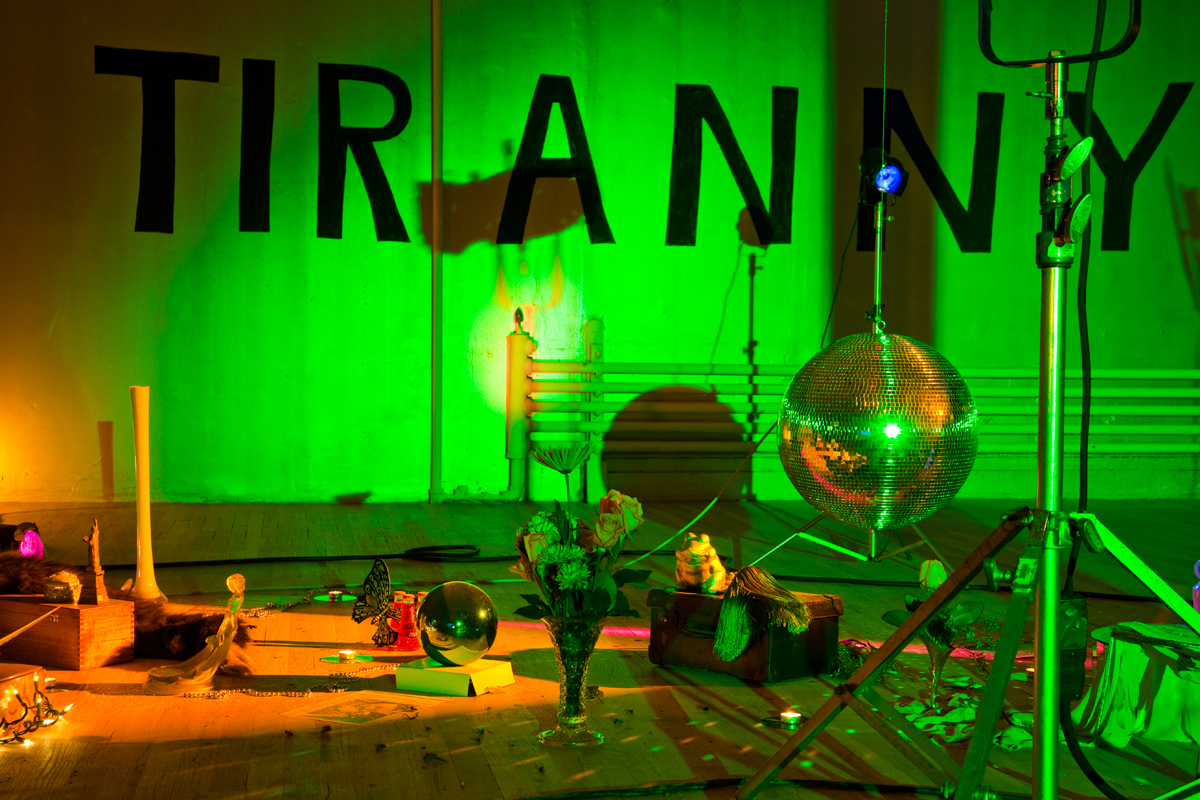 The word "TIRANNY" is handwritten in block letters on a wall. In front of it, there is a low-hanging disco ball, surrounded by various objects set on the floor, including a vase with flowers, a silver ball, a brown box, and more. The room is lit with a green hue.
