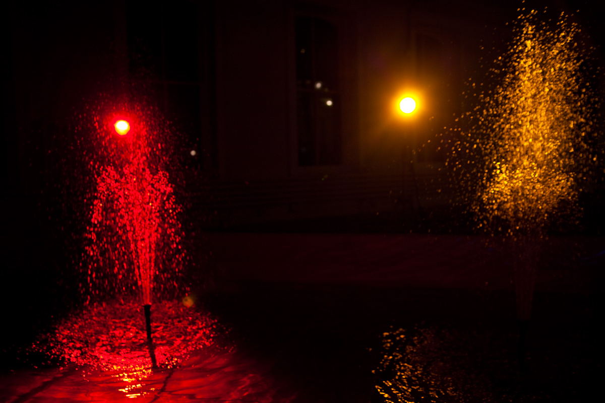 Two fountains of water in a dark room. The stream on the left is lit from behind by a red light, and the right stream by a yellow light.