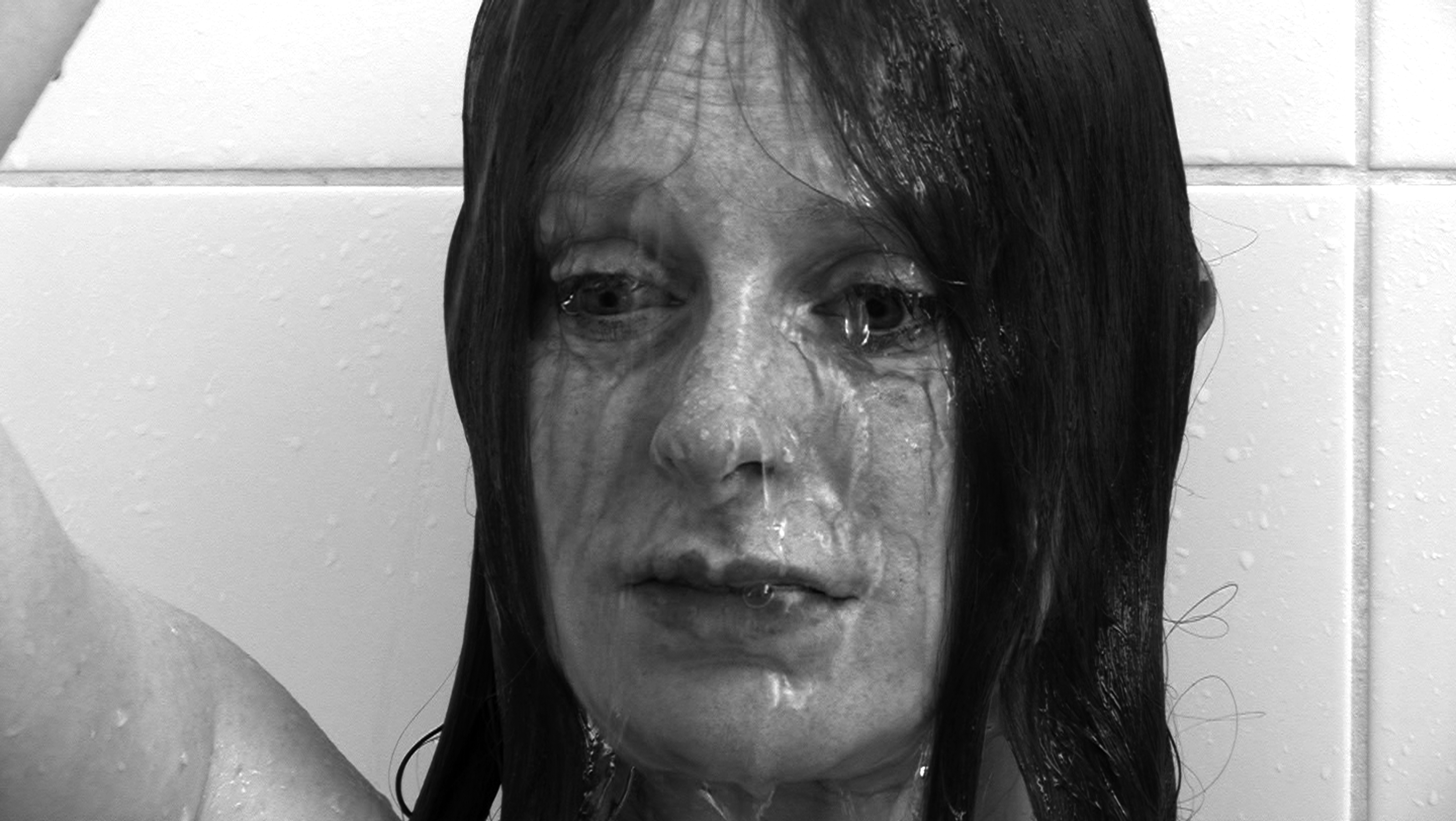 A black and white image of a person's face against a white tiled wall, with water pouring down over her.