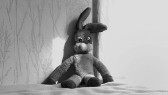 A black and white image of a floppy, stuffed rabbit toy. It is sitting on fabric and leaning up against a wall with faint floral design.