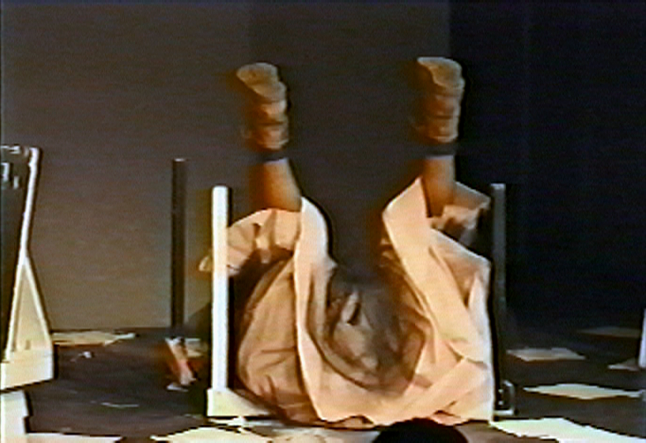 Video still of a figure laying on their back on top of an overturned, white table on a small stage area. They stretch their legs straight up above themselves. They wear a pink dress with black tulle fabric and brown boots. Scattered papers are strewn around them onstage.