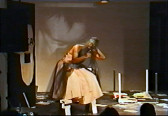 Video still of a figure in glasses wearing a pink dress with black tulle fabric on a small stage. An overturned, white table is siutated to their right, and papers are strewn across the ground. The figure hunches over, looking out towards the audience. Their left hand is bent at their chest and points upwards, while their right hand touches the back of their head.