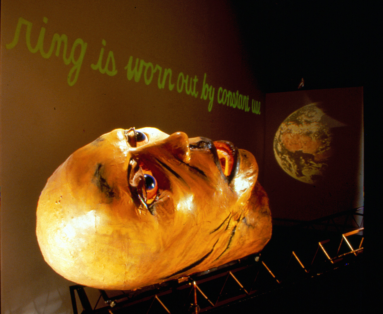 A large paper-maché head lays face-up on a metal scaffolding.  Above it, green cursive text reads, 