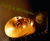 A large paper-maché head lays face-up on a metal scaffolding.  Above it, green cursive text reads, 