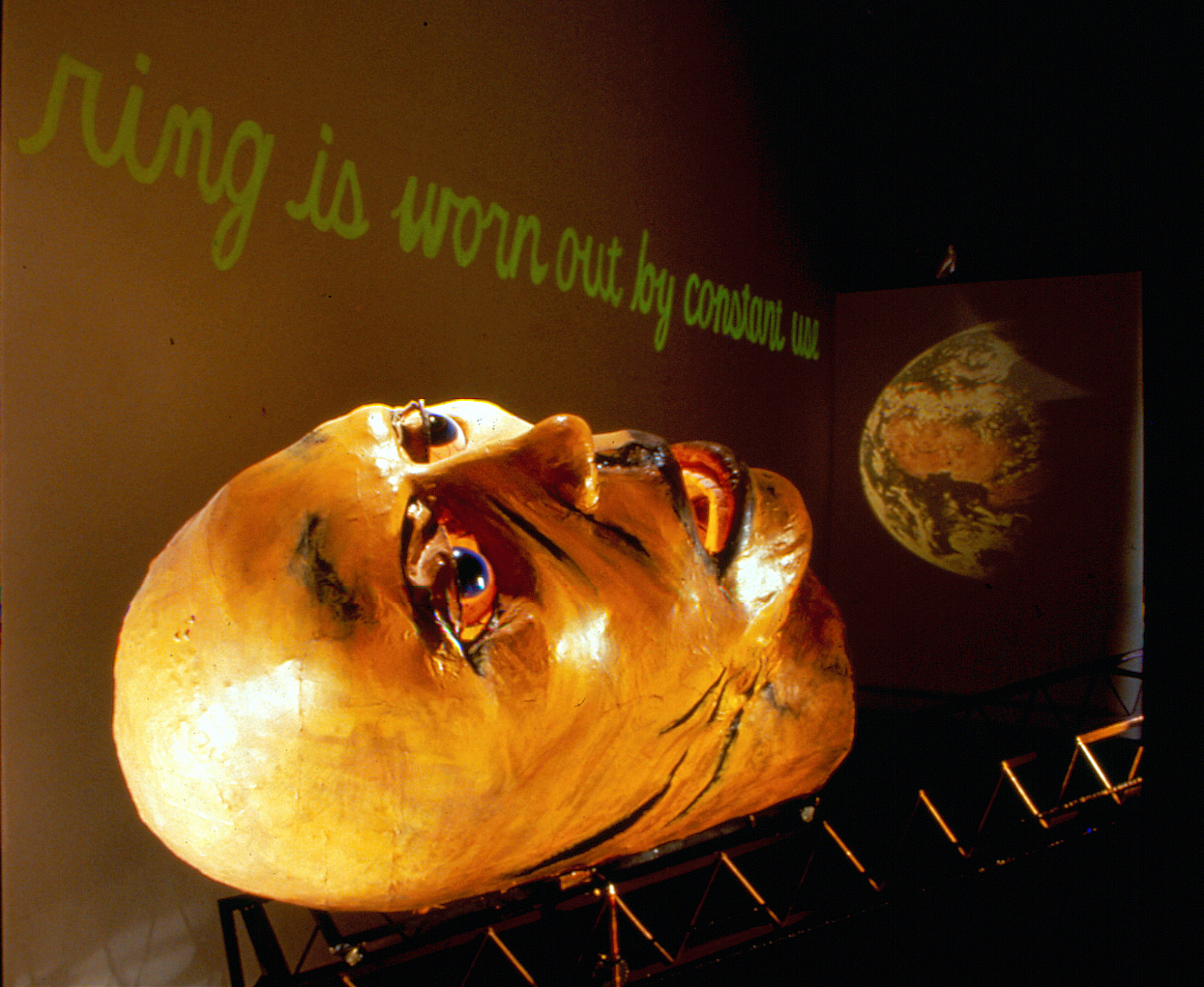 A large paper-maché head lays face-up on a metal scaffolding.  Above it, green cursive text reads, 