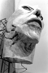 A large, hollow paper mache head is seen from below in a black and white photograph.  The head is held up by a metal armature and scaffold.