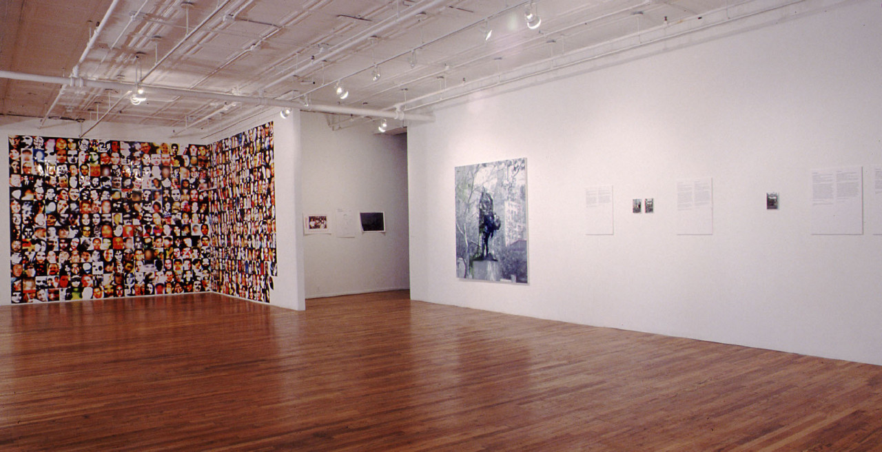 A large gallery space with a wooden floor and white walls. On one wall that forms an L shape into the space, slightly distorted color portraits are arranged floor to ceiling in a tight grid. On the other white wall is a large black and white photograph, with several other small photographs with accompanying text.