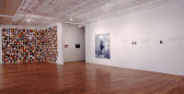 A large gallery space with a wooden floor and white walls. On one wall that forms an L shape into the space, slightly distorted color portraits are arranged floor to ceiling in a tight grid. On the other white wall is a large black and white photograph, with several other small photographs with accompanying text.