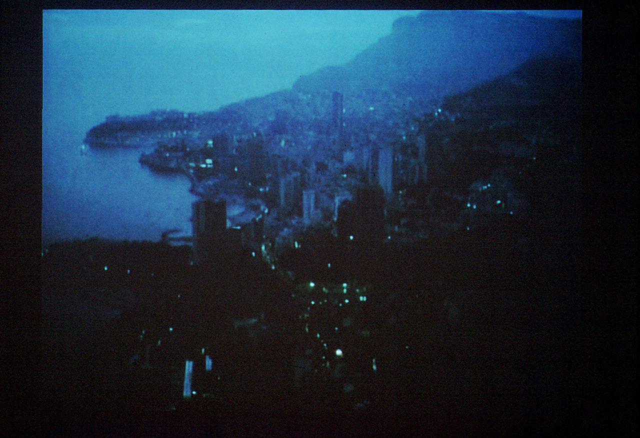 A dim, twilight image of a city on a coastline. Bright, turquoise blue streetlights dot the photograph.