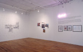 A large space with a wooden floor and white walls. On one wall is a black and white floorplan with 3D structures. On the other wall, there are a series of photographs with several small street signs arranged in a clump. To the right of that, there is a block of wall text with four frames below. Two of the frames hold *Time Magazine* covers.