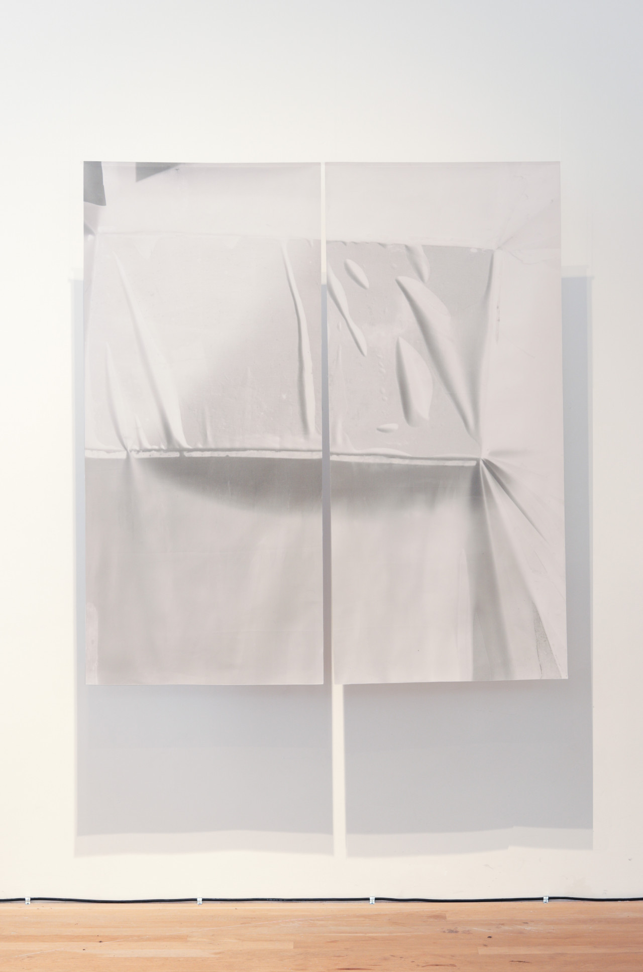 Color photograph of a work consisting of two large sheets of textile with shades of white and grey, suspended from the ceiling. The work casts a shadow on the wall behind it.