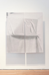 Color photograph of a work consisting of two large sheets of textile with shades of white and grey, suspended from the ceiling. The work casts a shadow on the wall behind it.