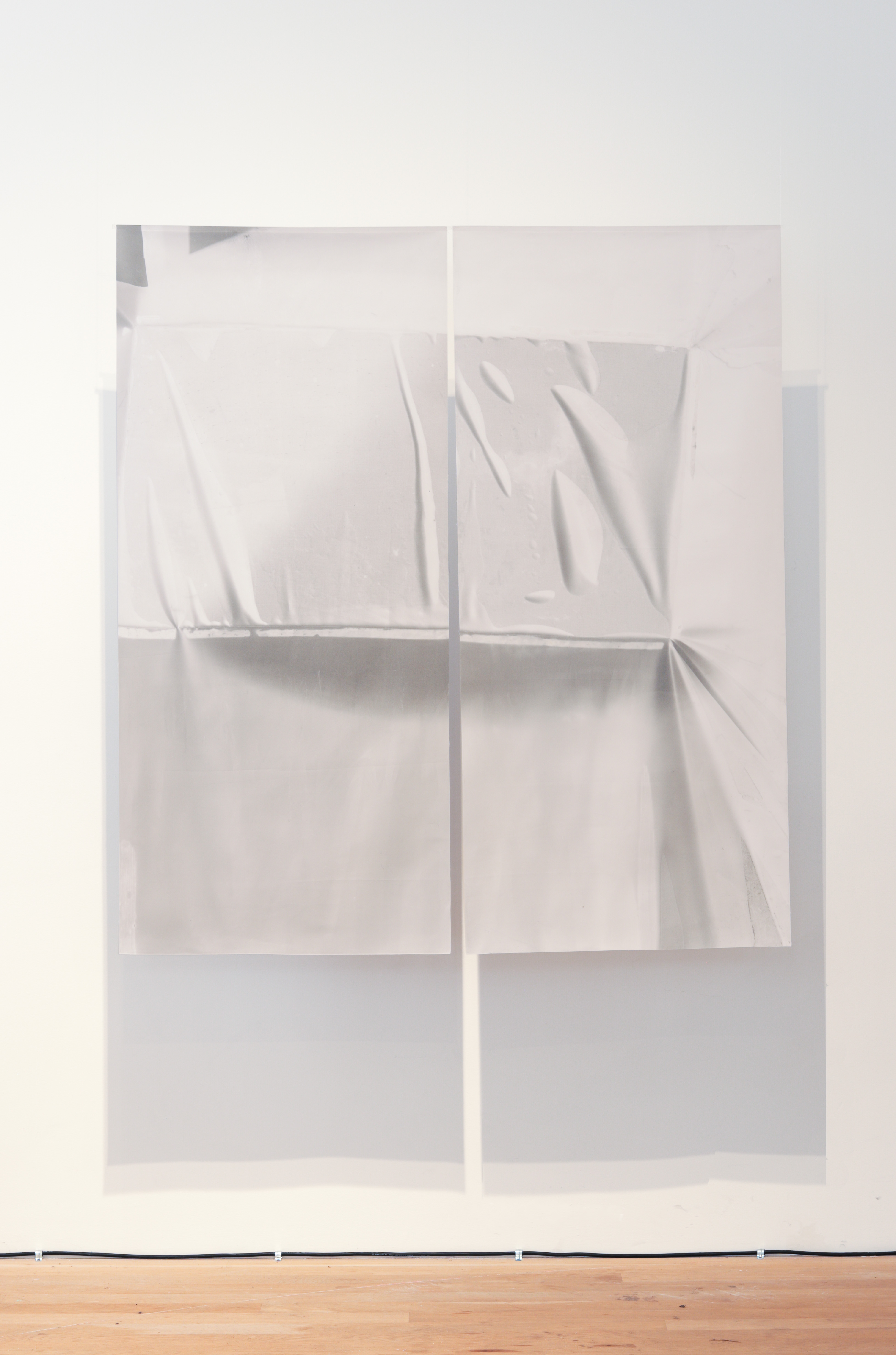 Color photograph of a work consisting of two large sheets of textile with shades of white and grey, suspended from the ceiling. The work casts a shadow on the wall behind it.