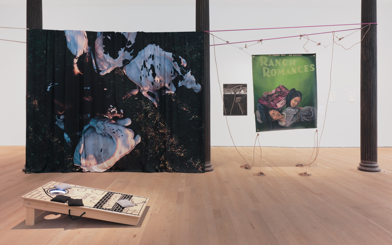 A color photograph of a gallery space with several columns. In between two columns, a large fabric work is stretched with an image of cows lying on the ground. To the right, in between two other columns, a painting on fabric that depicts two people on a green background is hanging. Further in the background, a painting is visible hanging on the wall. In the foreground, one board from a cornhole set is resting on the floor.