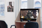Color photograph of video work installed on a wooden desk. Visible on the wall in the background is a work on paper mounted on a mirror.