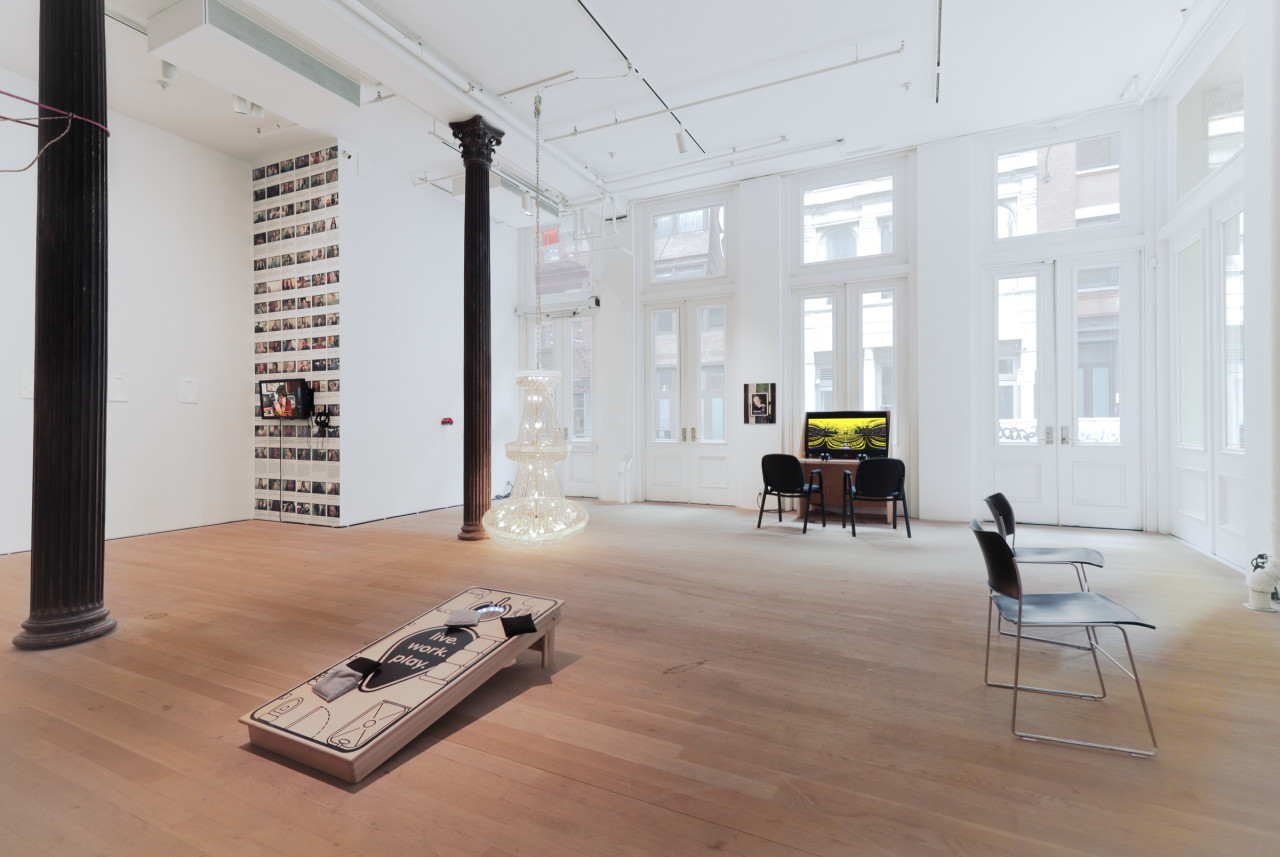 Color photograph of a gallery space with two dark columns. In the foreground, a cornhole board is visible resting on the floor. In the background, a work on a video monitor is installed on a desk that has two chairs in front of it. A chandelier hangs from the ceiling near the furthermost column and rests on the floor. In the left side of the background, a wall is filled with images and has a mounted monitor with a video work.