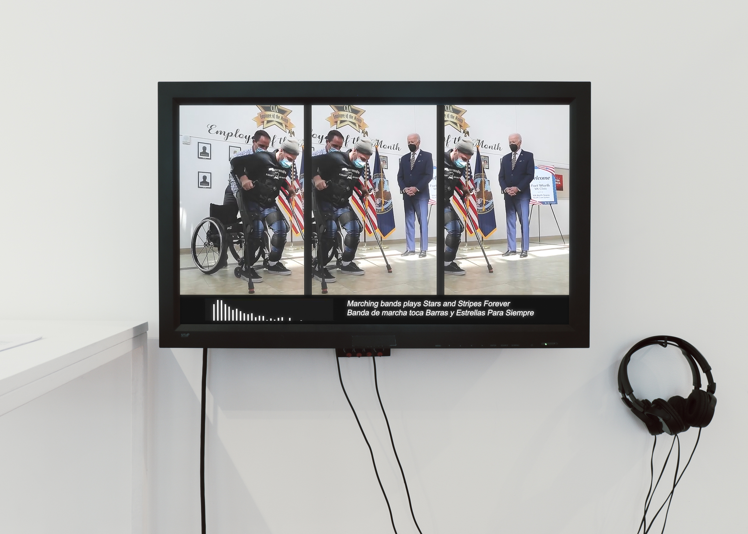 A color photograph of a video monitor mounted on a wall with two headsets hanging nearby