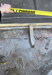 A person holding caution tape over a patch of wet cement with the words <i>Everything is common</i> written in it.