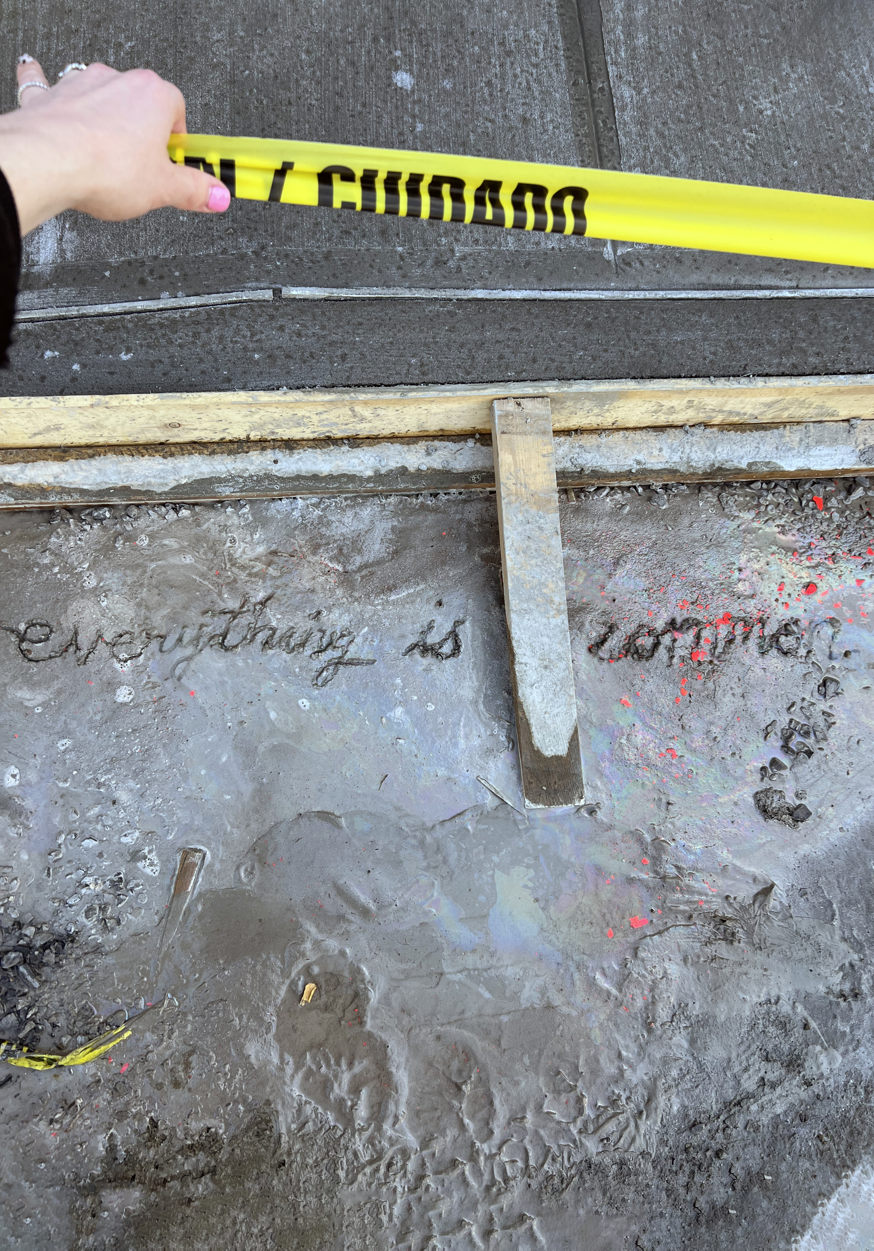 A person holding caution tape over a patch of wet cement with the words <i>Everything is common</i> written in it.