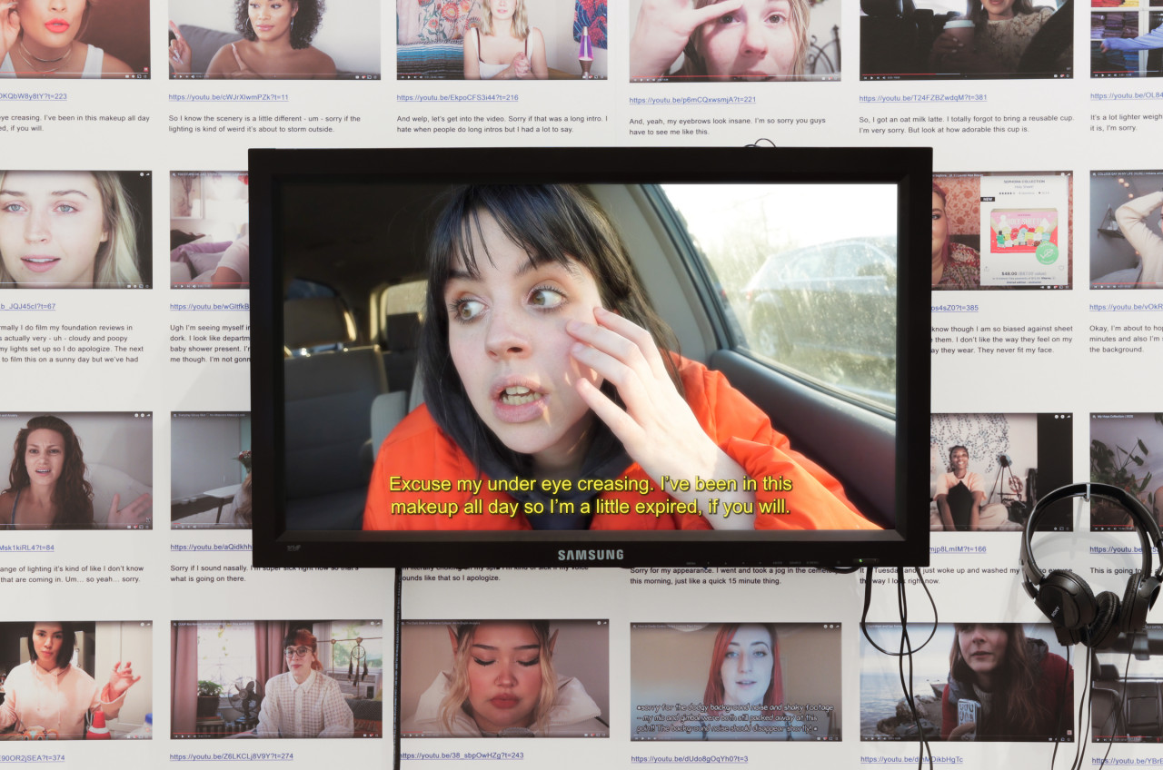 Color installation image showing a video monitor mounted on a wall which is covered in tiled images of YouTube pages. A pair of wired headphones is hanging to the right of the monitor.