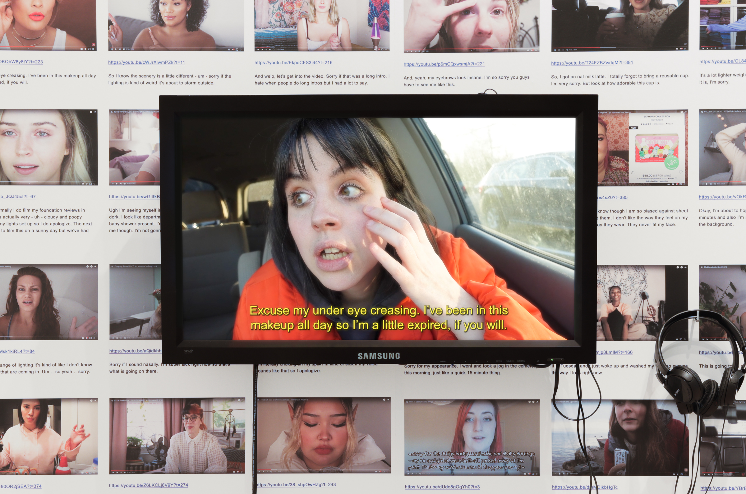 Color installation image showing a video monitor mounted on a wall which is covered in tiled images of YouTube pages. A pair of wired headphones is hanging to the right of the monitor.