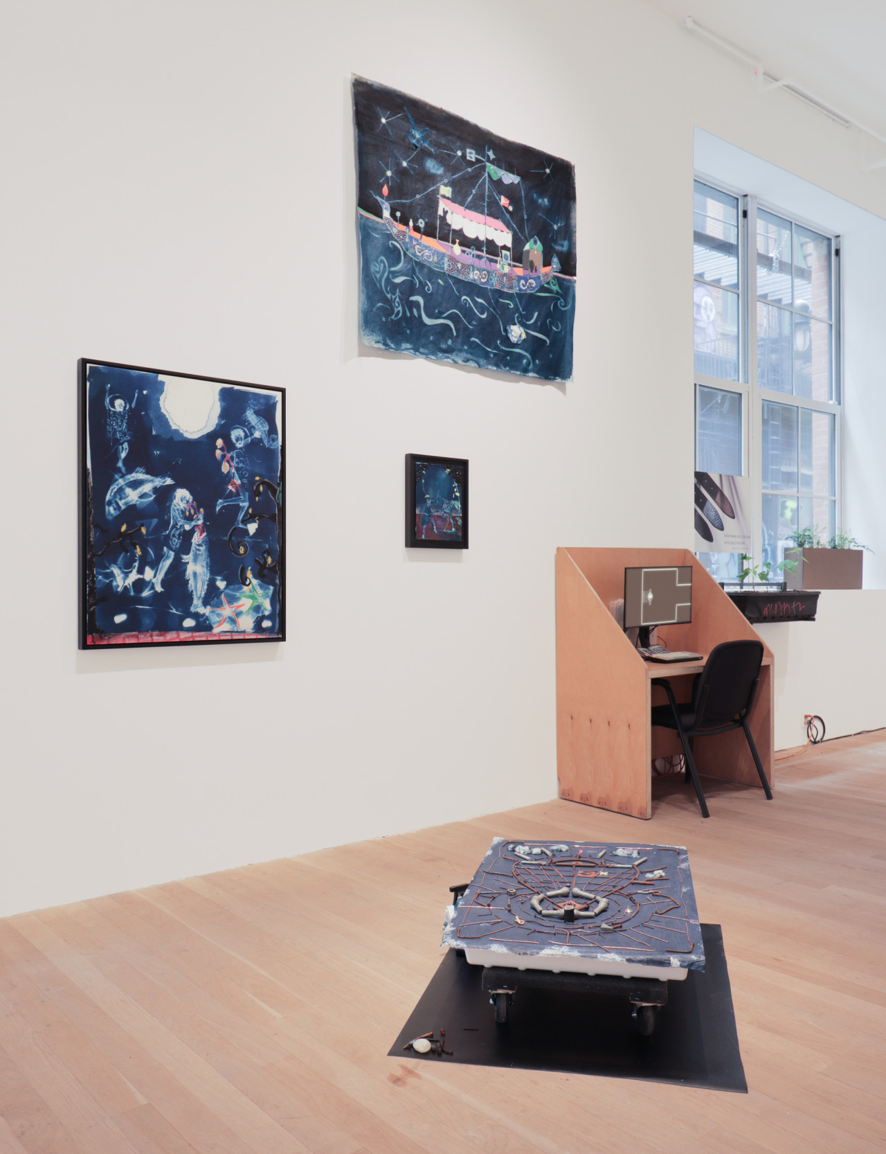 A color photograph of a gallery space. In the foreground, a work slightly elevated from the ground has several objects resting on its surface. On the background wall two larger works on paper and one smaller one are hanging. A desk with a video work on a monitor is positioned against the wall.