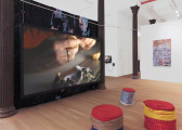 A color photograph of a galllery space with several dark columns. In between two columns in the foreground is a large fabric projector screen, with a video being projected onto it. In front of the screen are several fabric-wrapped stools. Further in the background a painting is visible on one wall, and a tv screen with a video work is visible on another.