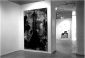 In the foreground is one dark painting image and in the background is a painting of a figure coming out of a square brick chimney-like structure with marking above them. These two paintings appear in a white-walled gallery space.