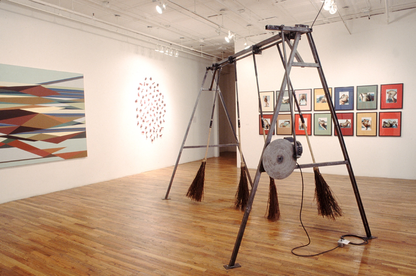 A gallery space with a large metal structure in the middle of the floor. Four straw brooms are suspended from the top of the structure such that they just brush the floor. On the surrounding white walls, there are several pieces of art. One is a colorful canvas with geometric shapes, sharing a wall with several small objects arranged in an oval. On the other wall, there are series of framed depictions of nude figures in various poses.