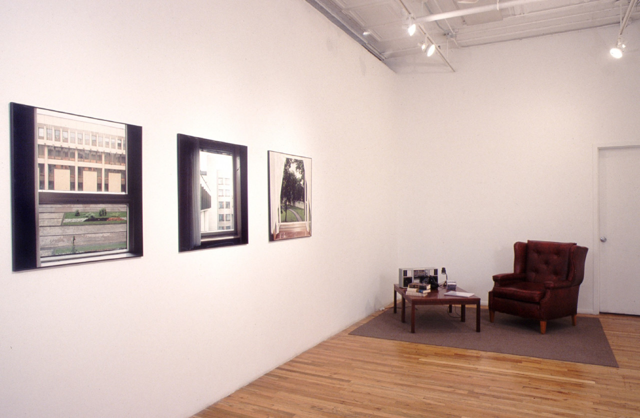 A red leather armchair with a coffee table on a gray rug in the corner of a room. On the coffee table, there are several books, a lamp, and an old radio. On the opposite wall, there are three photographs each showing the view out of three different windows.
