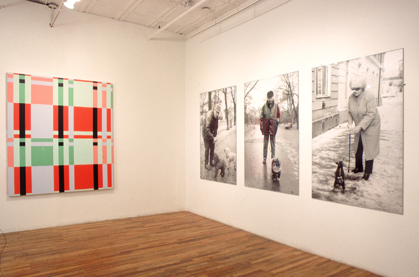 Two white walls meeting in a corner. The wall on the right has three large black and white photographs of people on winter streets with their small dogs. The wall on the left features a canvas covered in a red, green, and peach colored plaid pattern.