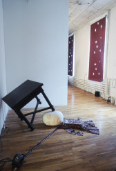 A small black table leans at an angle, one corner resting on a white wall and one leg resting on an inflated balloon with a face printed on it. Coming out of the ballon is a flowered dress, attached to a fan by an inflation tube. Beyond that, visible behind a half wall, are two red shades with holes covering two windows.