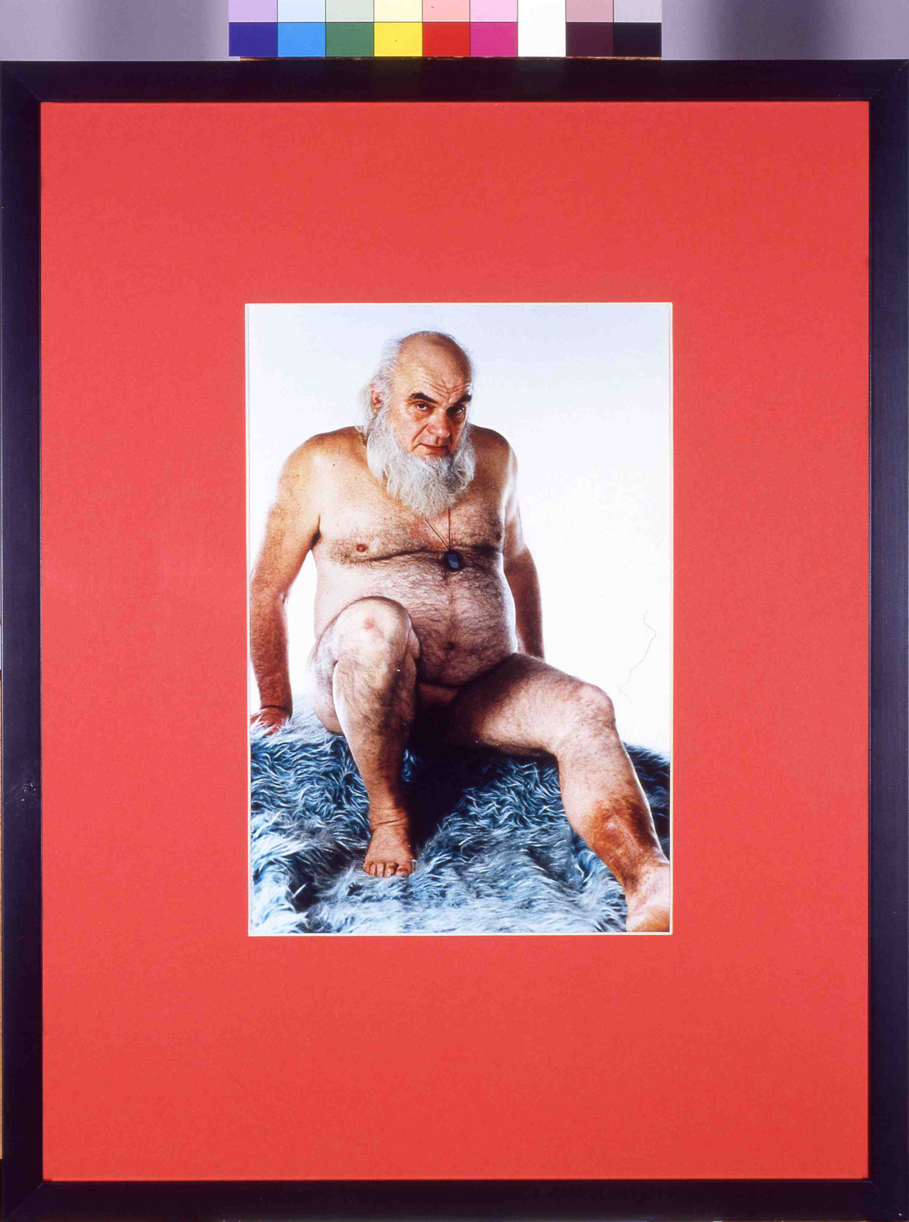 A framed photograph with a red border of a naked man on a blue shag carpet. The man is seated, leaning slightly back on his hands with his legs extended. He is bald with a full grey beard.