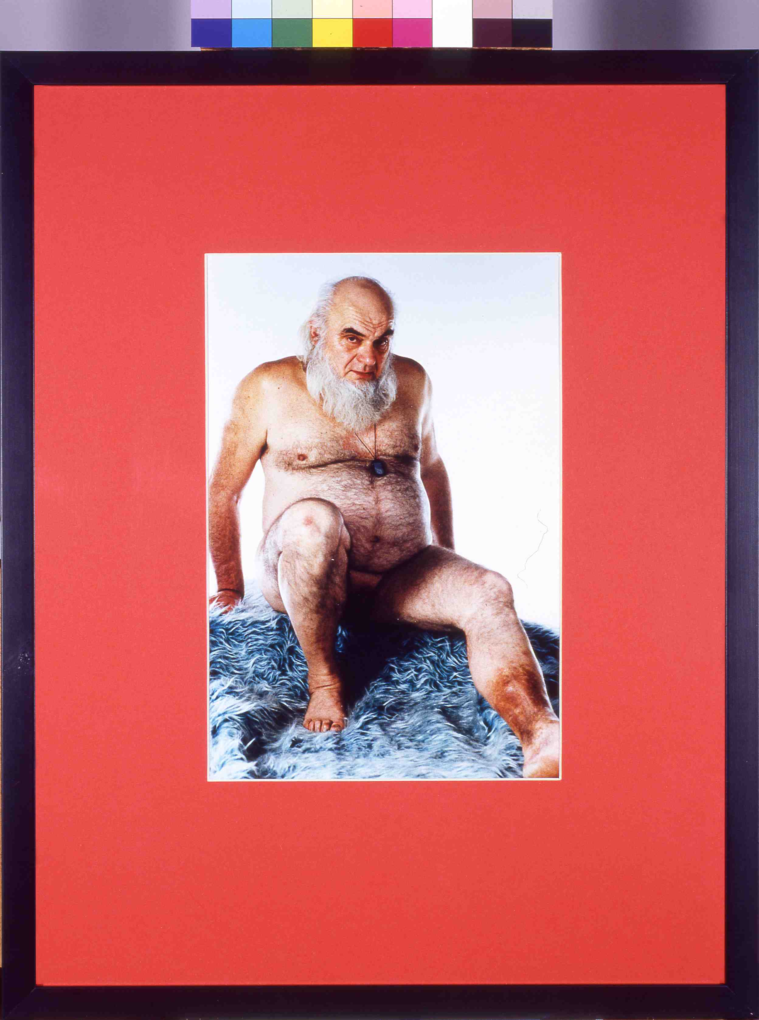 A framed photograph with a red border of a naked man on a blue shag carpet. The man is seated, leaning slightly back on his hands with his legs extended. He is bald with a full grey beard.