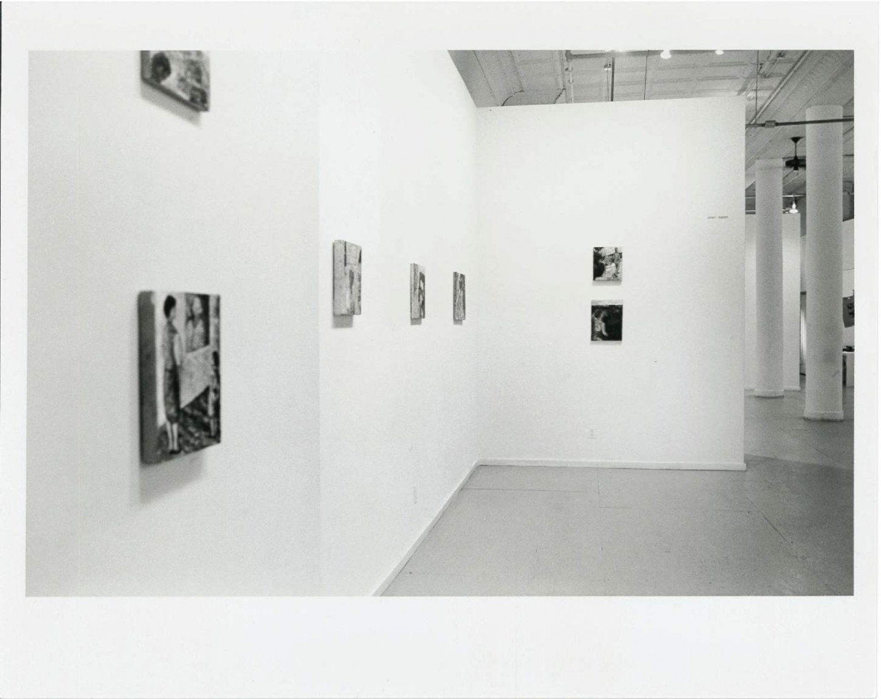 Paintings are hanging on walls in a white-walled gallery space with columns in the background.