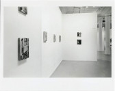 Paintings are hanging on walls in a white-walled gallery space with columns in the background.