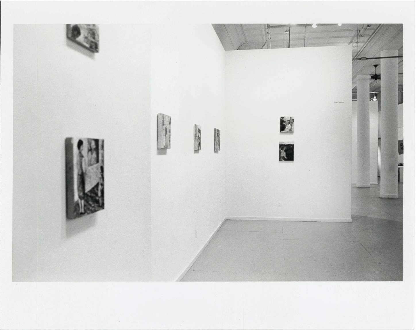 Paintings are hanging on walls in a white-walled gallery space with columns in the background.