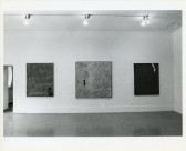 3 large canvases hang on a white wall. In the lower right-hand corner of the canvas to the left is a small image of a dog. On the left side of the center canvas is small a human figure. On the upper right-hand corner of the canvas to the right is a small image of a woman's ankle and foot.
