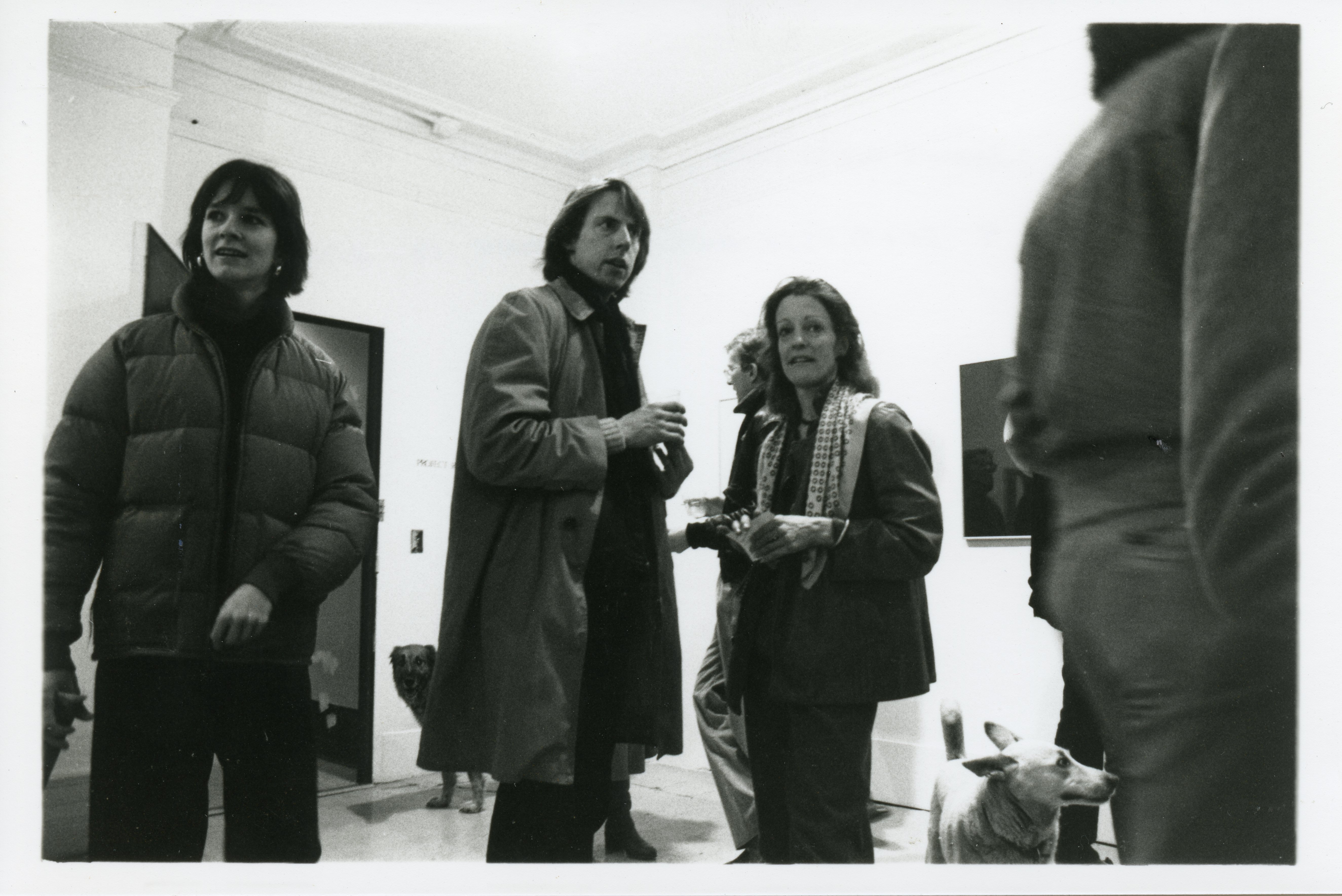 A photograph of the exhibition opening reception. A group of visitors look around at the art. Two dogs walk through the crowd.