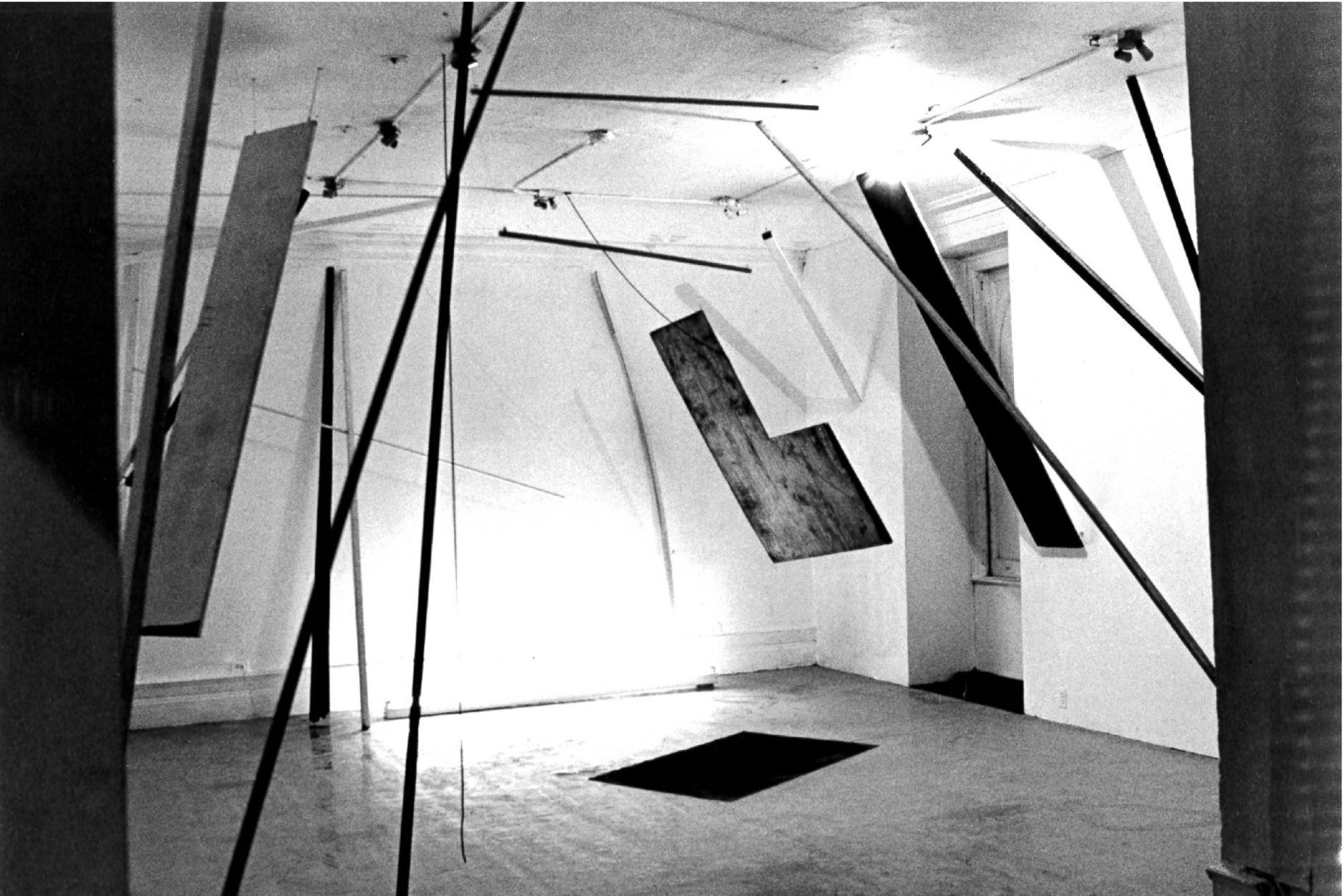 A black and white photograph of various objects hanging in a white-walled gallery space with a spotlight in center of the image.