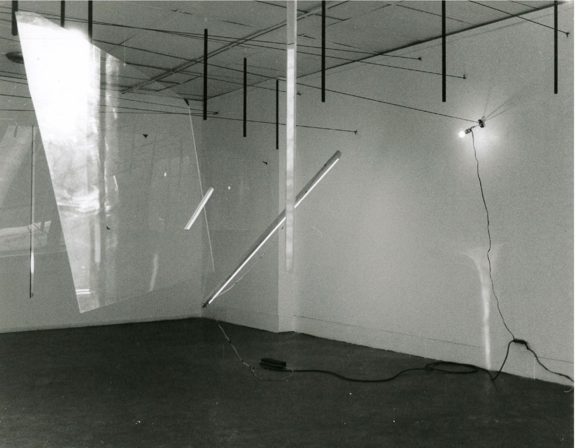 A black and white photograph of various objects hanging in a white-walled gallery space.