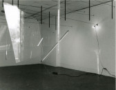 A black and white photograph of various objects hanging in a white-walled gallery space.