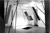 A black and white photograph of various objects hanging in a white-walled gallery space with a spotlight in center of the image.