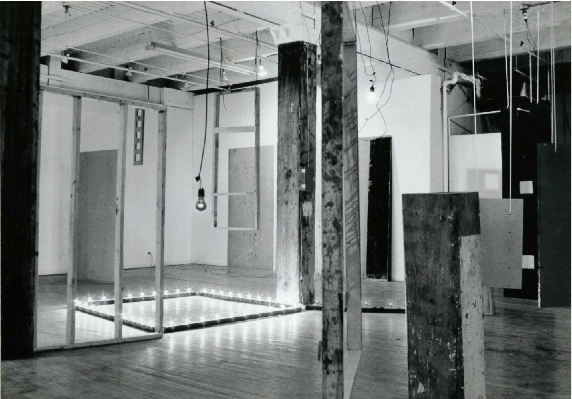 A black and white photograph of various objects hanging and standing in a white-walled gallery space.