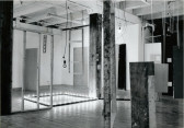 A black and white photograph of various objects hanging and standing in a white-walled gallery space.