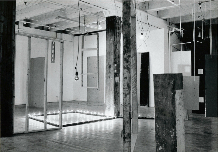A black and white photograph of various objects hanging and standing in a white-walled gallery space.