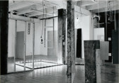 A black and white photograph of various objects hanging and standing in a white-walled gallery space.