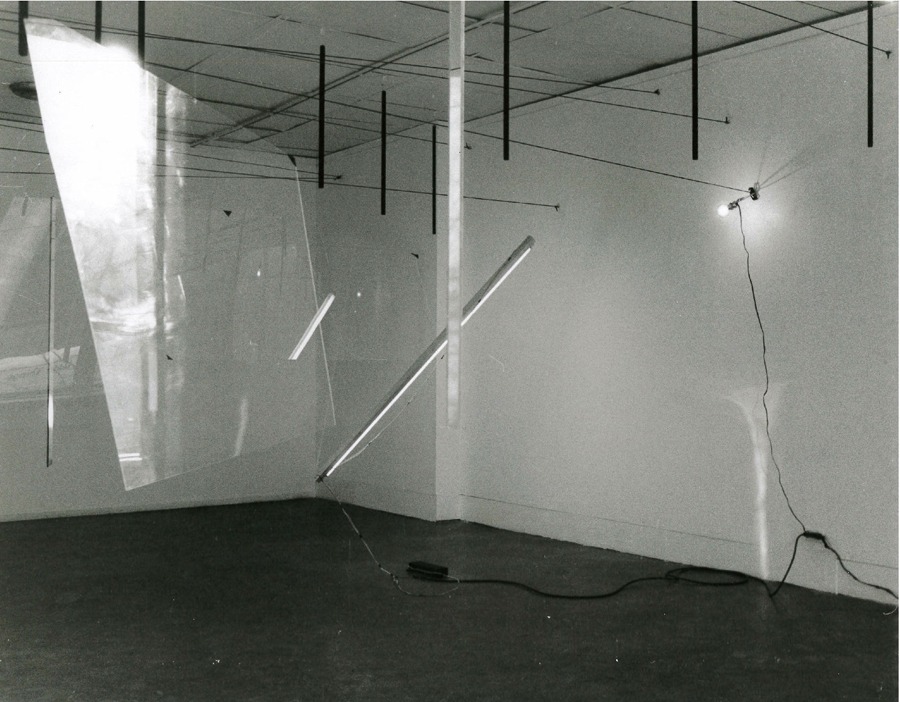 A black and white photograph of various objects hanging in a white-walled gallery space.