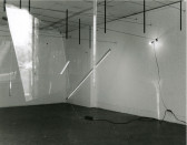 A black and white photograph of various objects hanging in a white-walled gallery space.